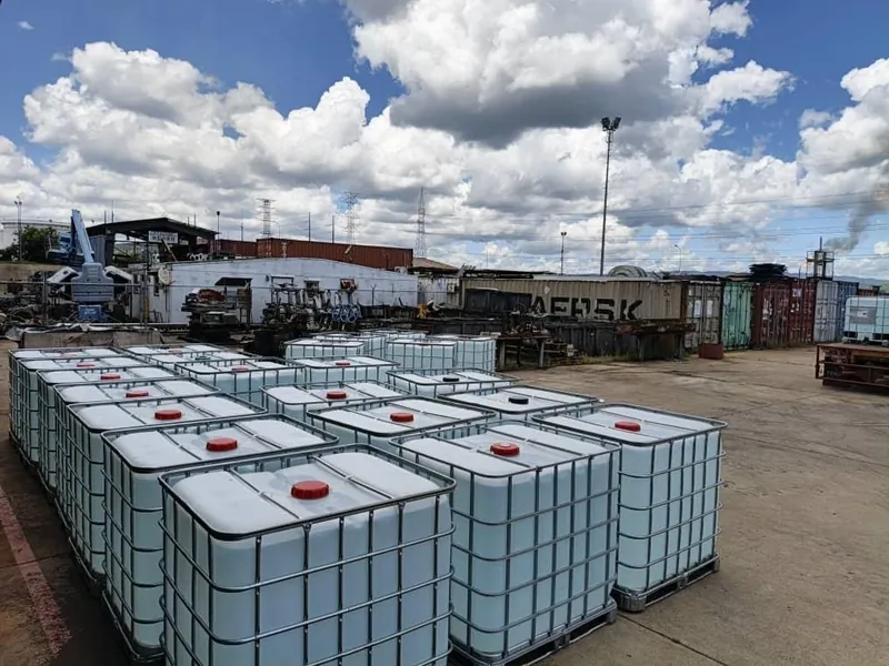 Filas de contenedores IBC blancos con tapas rojas sobre un patio de concreto bajo un cielo con nubes